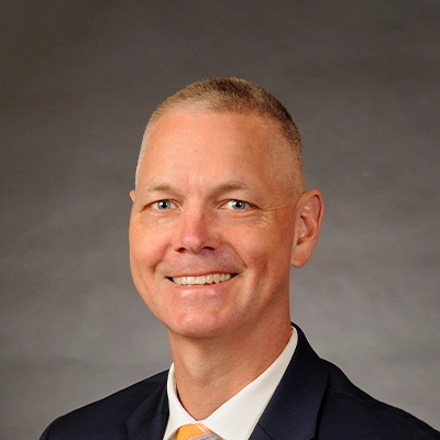 Chiropractor Todd Tesch Smiling In Black Suit In Front Of Grey Background